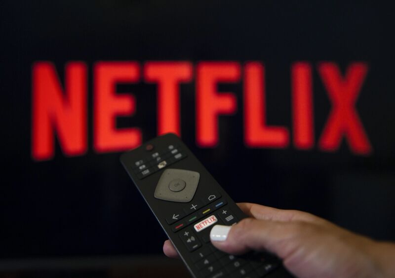 Netflix logosunu gösteren bir ekranın önünde uzaktan kumanda tutan kişi.