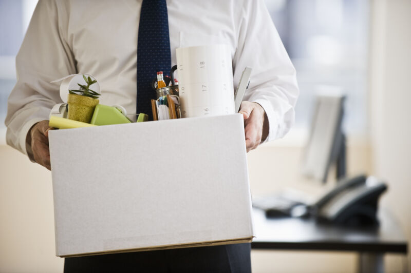 A man in an office walks away from his desk carrying a box of knickknacks.