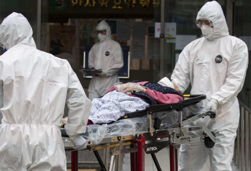 People in protective gear wheel a patient on a gurney.