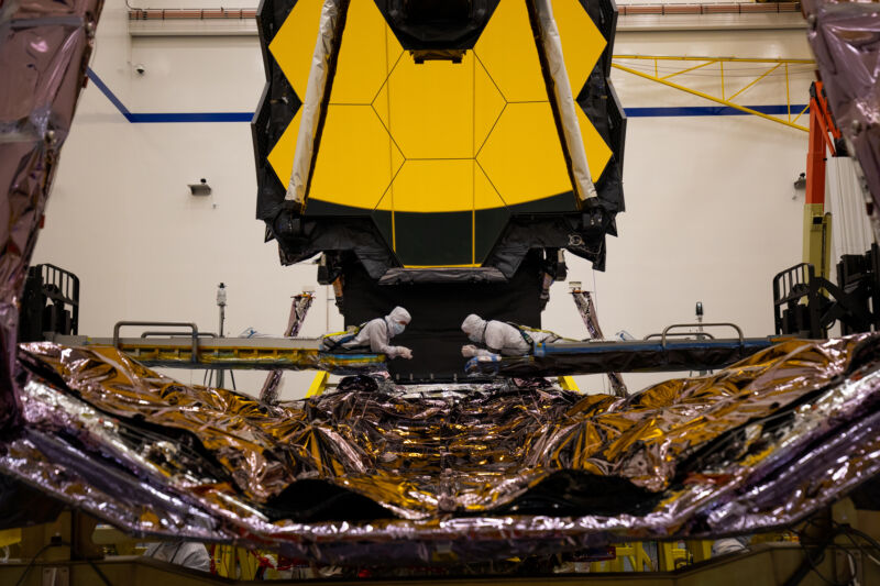 Two people in clean suits work inspect a huge satellite.