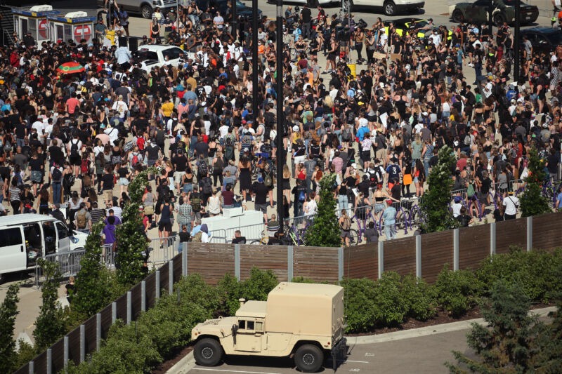 A crowd stands on one side of a fence from a tan-colored truck.