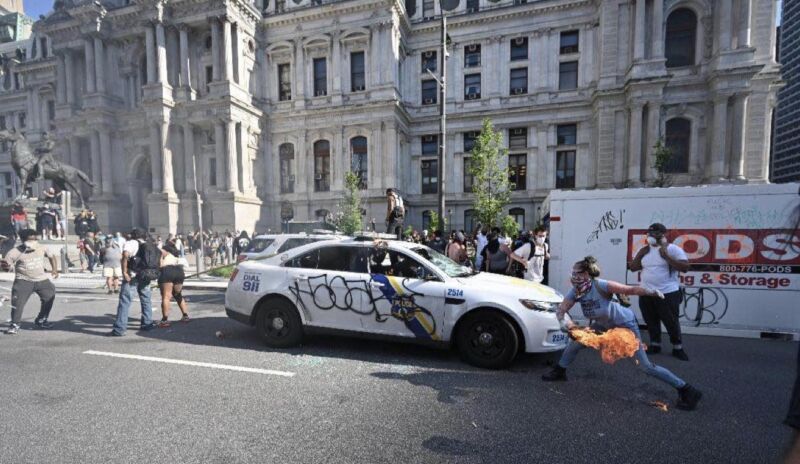 A masked woman holding a flaming piece of wood next to a police car.