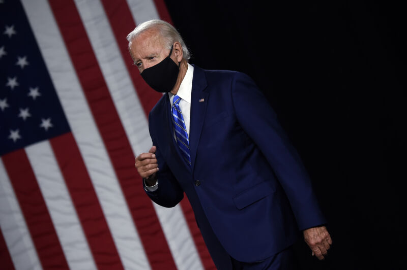 A older man in a suit and face mask walks past an American flag.