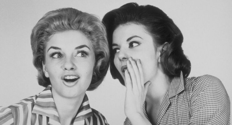 Black-and-white photo of two 1950s style women whispering.