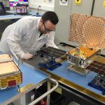 Engineer Joel Steinkraus stands with both of the MarCO spacecraft. The one on the left is folded up the way it will be stowed on its rocket; the one on the right has its solar panels fully deployed, along with its high-gain antenna on top.
