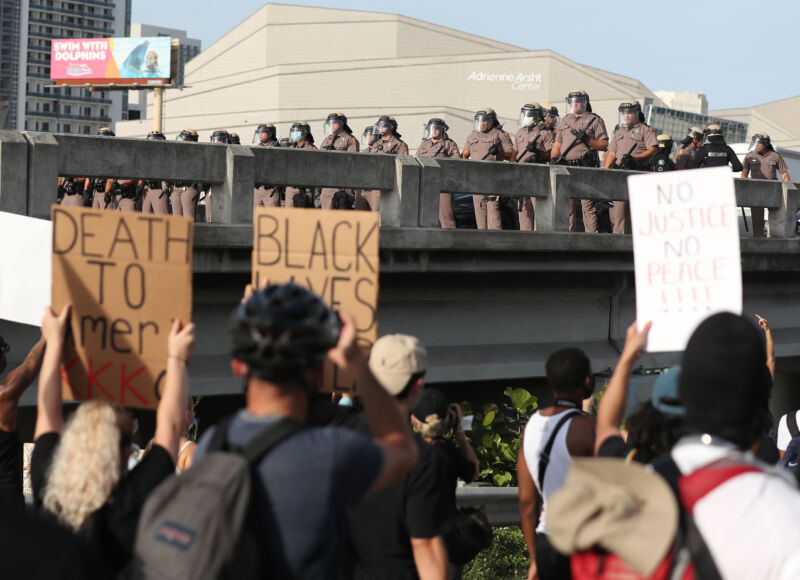 People hold up signs while police in riot gear watch from above.