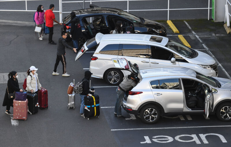 Masked travelers stand near cars parked in spaces set aside for Uber.