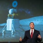 A man in a suit speaks in front of a mural of the Moon landing.