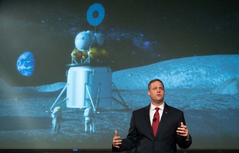 A man in a suit speaks in front of a mural of the Moon landing.