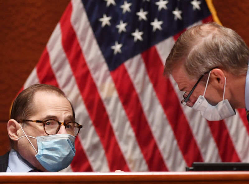 Two men in suits and face masks confer in front of a US flag.