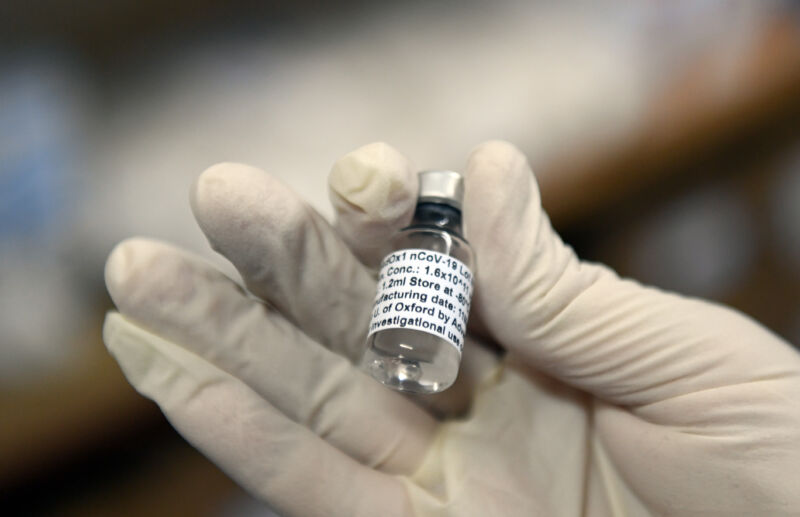 Close-Up Photograph Of A Gloved Hand Holding A Tiny Bottle Of Clear Liquid.