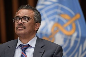 A man with a loosened necktie stands in front of a logo for the World Health Organization.