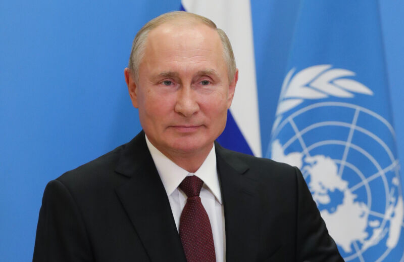 A smirking man in a suit sits in front of a UN flag.