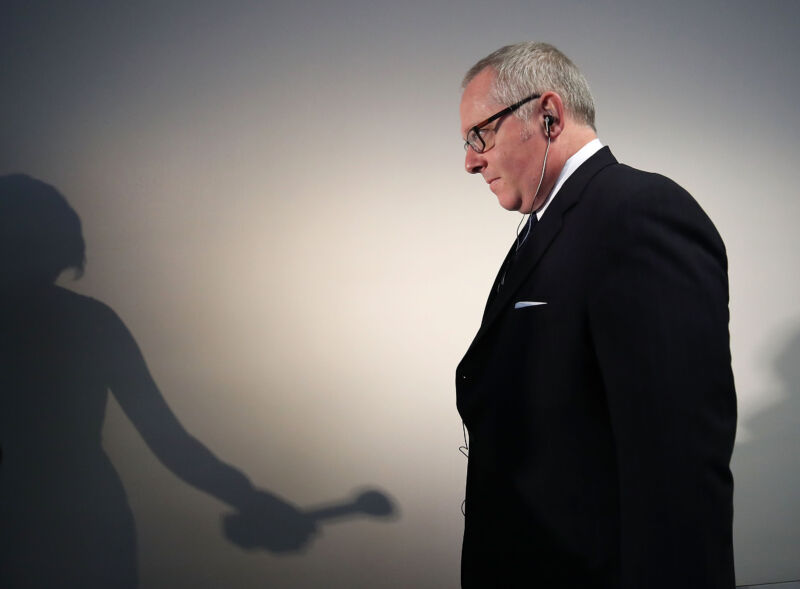 The shadow of a reporter with a microphone falls on the wall behind a man in a suit.