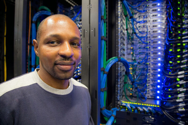 A casually dressed man smiles next to exposed computer components.