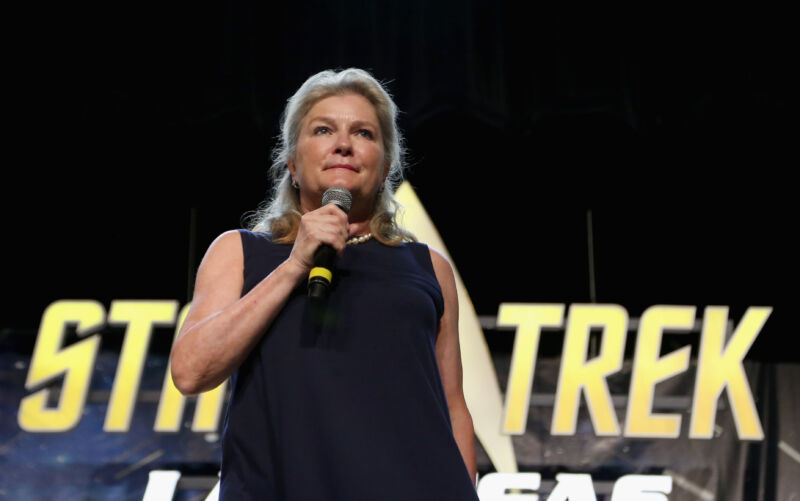 A woman speaks into a microphone in front of the original Star Trek logo.