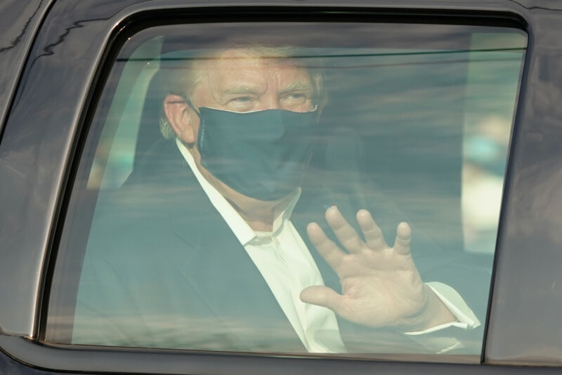 A masked man waves through a bulletproof window.