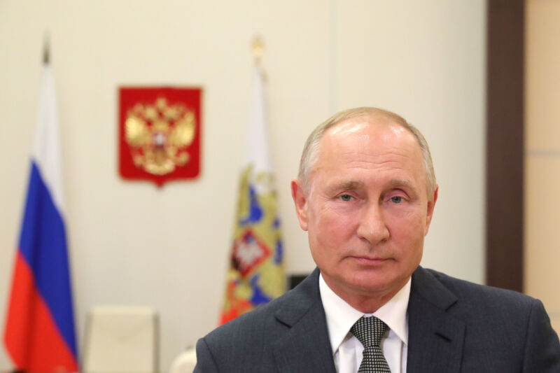 A vaguely smug man in a suit sits in front of a flag.