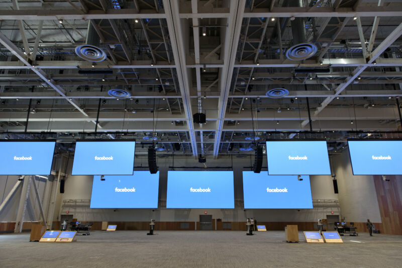 Giant monitors displaying the Facebook logo hang from the ceiling of an empty convention center.