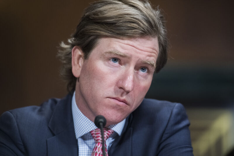 A somewhat irritated-looking man in a suit listens from behind a microphone.