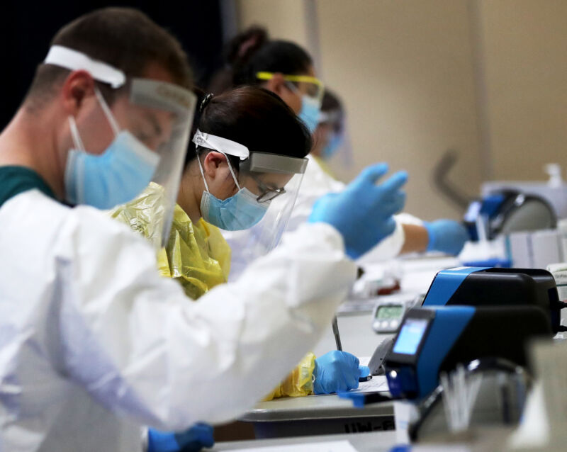 Workers in full gowns, masks, face shields, and gloves work at a table to process tests for COVID-19