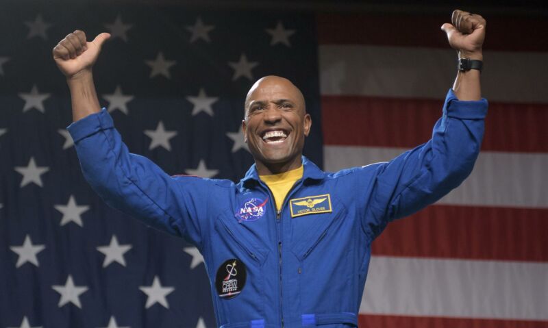 A man in a NASA jumpsuit strikes a victorious pose in front of a US flag.