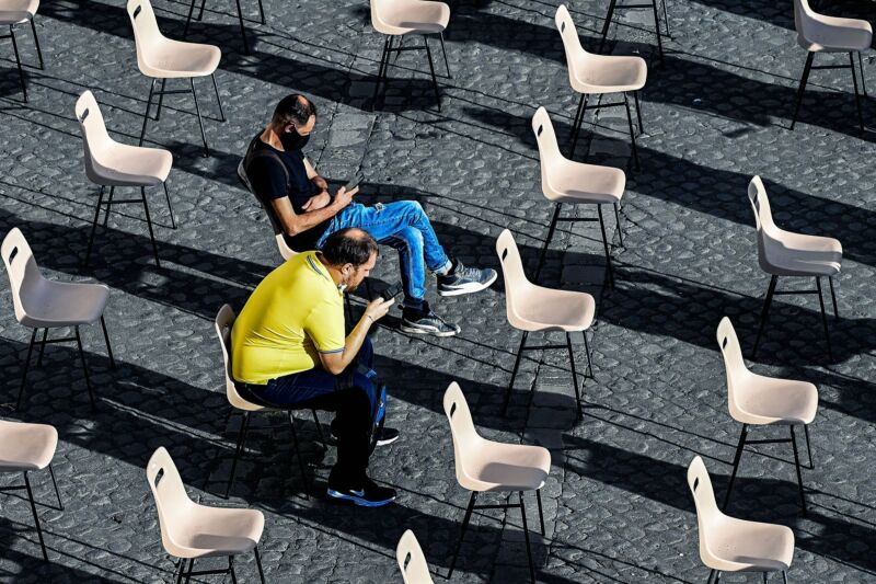 Several Rows Of Outdoor Seating Are Empty, Save Two Men Staring At Their Smartphones.