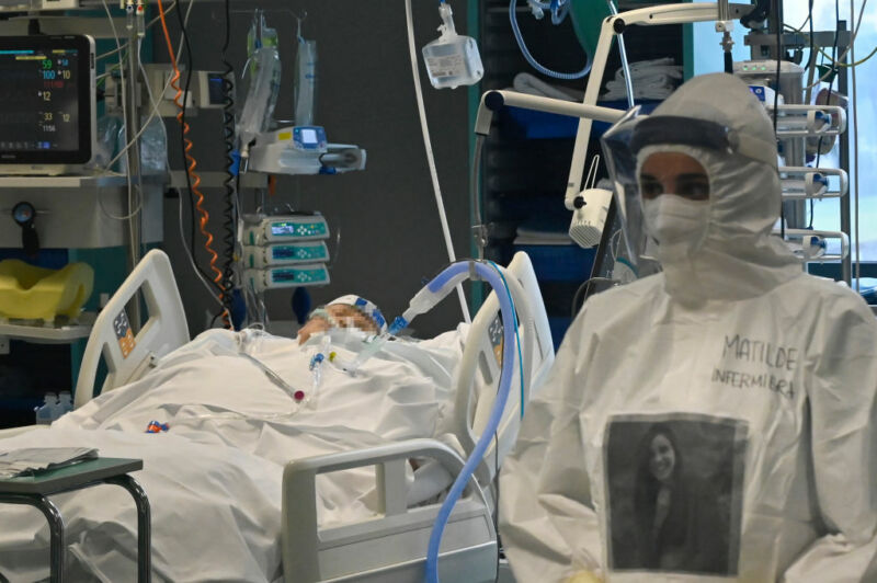 A medical worker in protective gear stands beside a bedridden patient hooked into all manner of machines.