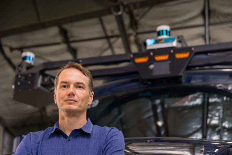 A casually dressed man stands in front of a large automobile.