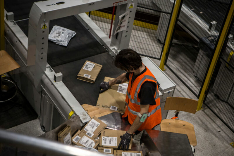 Un hombre con un chaleco naranja mueve paquetes de Amazon en una cinta transportadora.