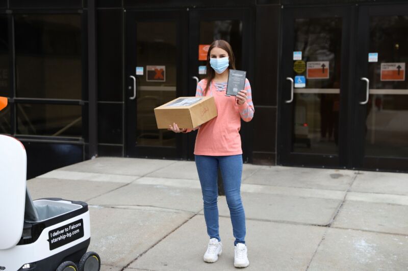A masked woman holds up a package next to a self-driving robot about the size of an ice cooler.
