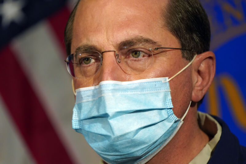 A serious man in glasses and a face mask addresses a crowd.