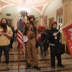 Men with flags and bizarre costumes pose for a photo in a neoclassical corridor.
