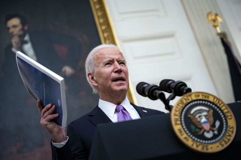 A Man In A Suit Holds Up A Laminated Binder While Speaking At A Microphone.