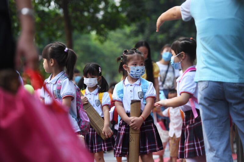 Masked girls in matching uniforms wait for school to begin.