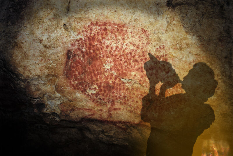 Color photo of a person with a conch shell raised to their mouth, silhouetted against a red-painted cave wall.