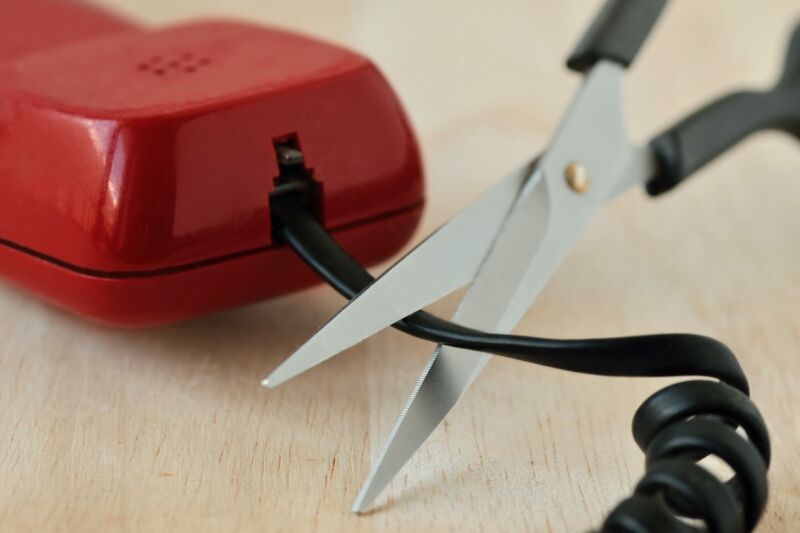 A pair of scissors being used to cut a wire coming out of a landline telephone.