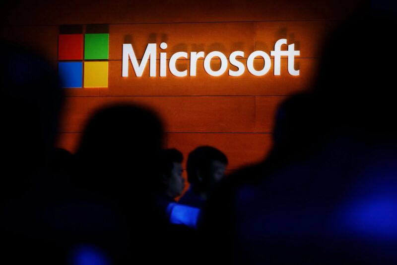 Gölgeli figürler, suni ahşap bir duvarda bir Microsoft logosunun altında duruyor.