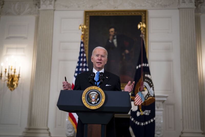 An elderly man in a suit speaks on a stage in front of a portrait of the Great Emancipator.