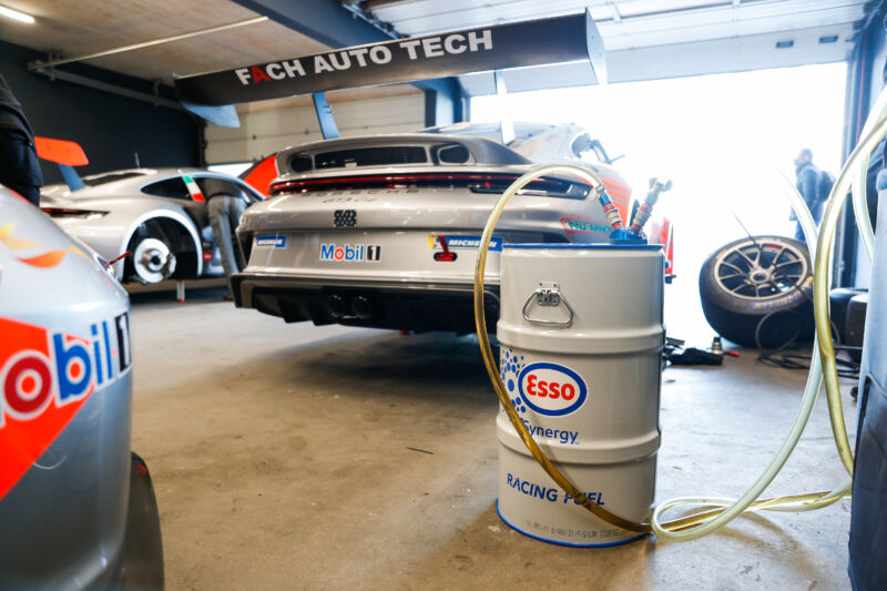 A barrel of fuel in front of a Porsche racing car in its garage.