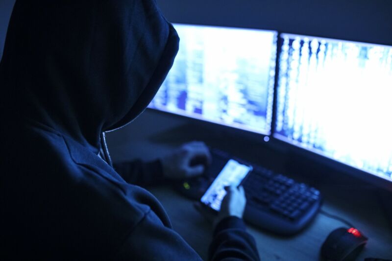 A hacker sitting in front of two computer screens and holding a smartphone.