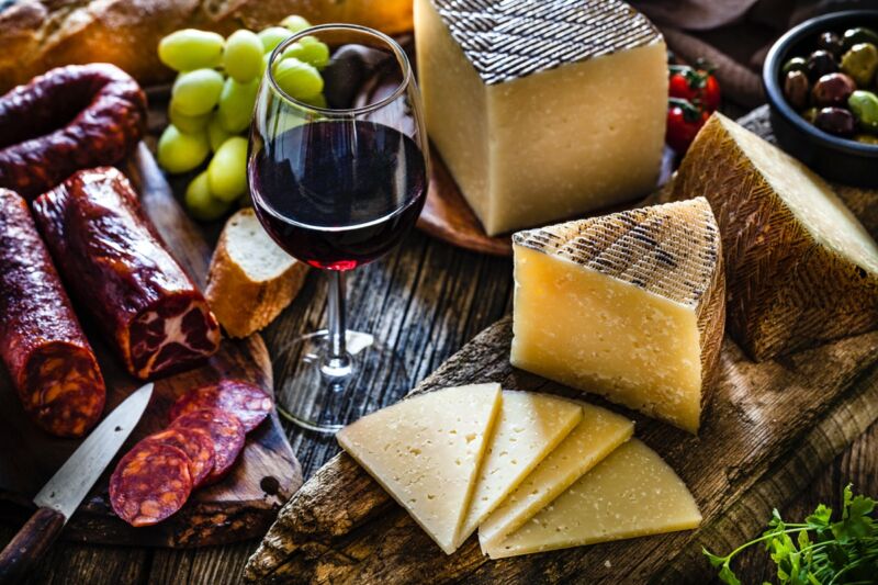 Tranches de fromage Manchego et coupées dans une planche à découper collée sur une table en bois sombre rustique, avec un verre de vin rouge.