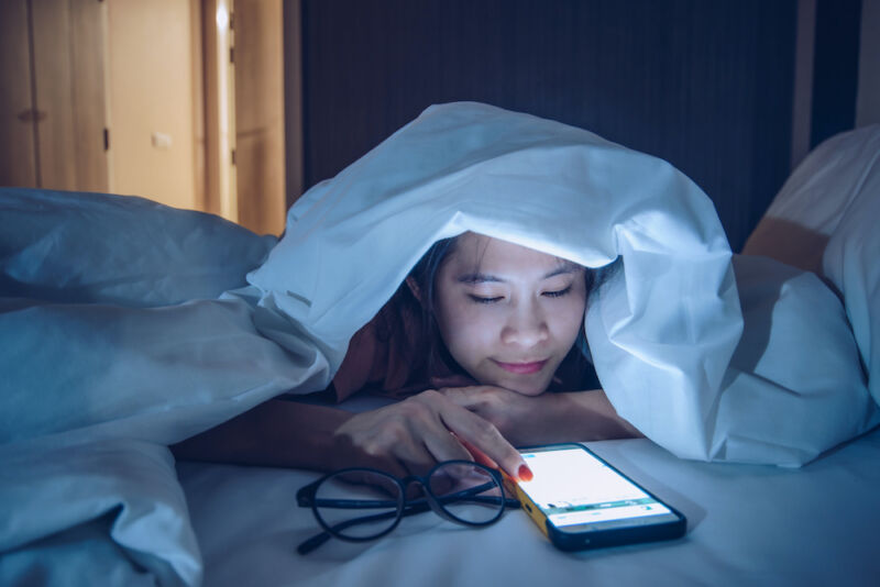 Una mujer debajo de una manta gruesa mira su teléfono inteligente.