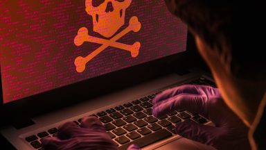 Gloved hands manipulate a laptop with a skull and crossbones on the display.