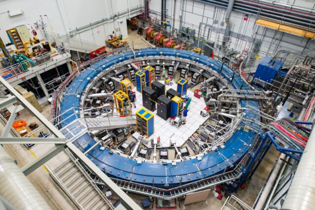 The Muon g-2 ring sits in its detector hall amid electronics racks, the muon beamline, and other equipment. This experiment studies the precession (or wobble) of muons as they travel through the magnetic field.