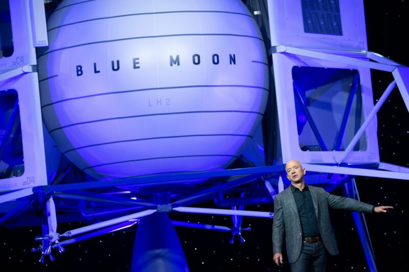 A man in a sport coat speaks next to a life-sized image of a small spacecraft.