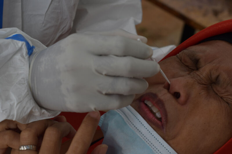 A gloved hand rams a cotton swab into an unhappy person's face hole.