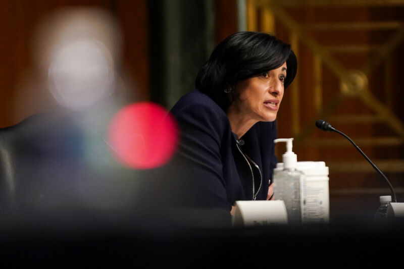 A serious woman speaks into a microphone while seated next to a pump dispenser of hand sanitizer.