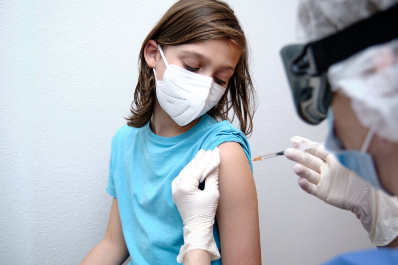 A masked child watches a healthcare worker perform an injection.