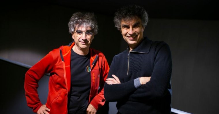 Two casually dressed men with tousled, salt and pepper hair pose for a photo.
